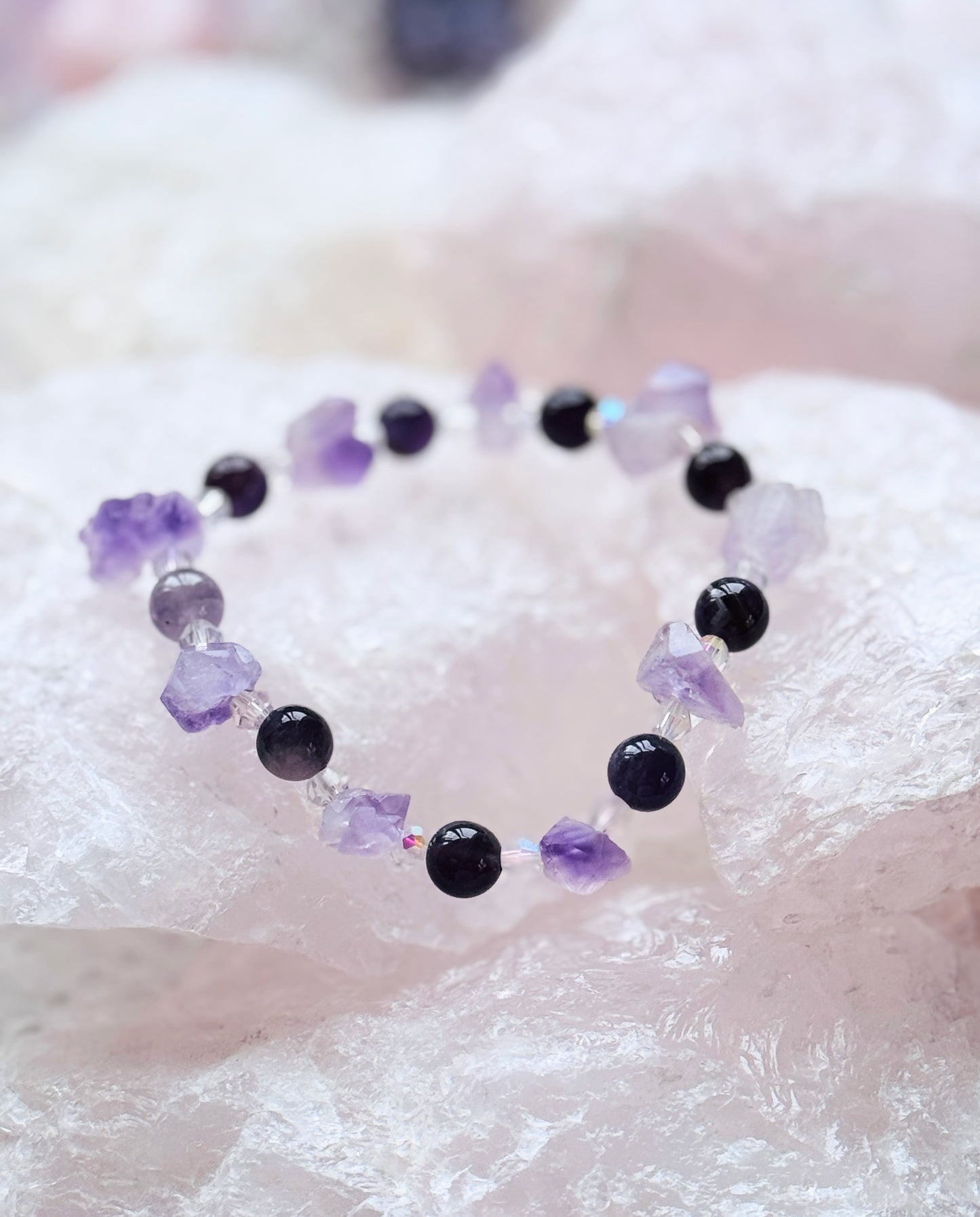 Crystal bracelet with amethyst flower stones and dark beads on pink crystal background, offering calm and clarity.