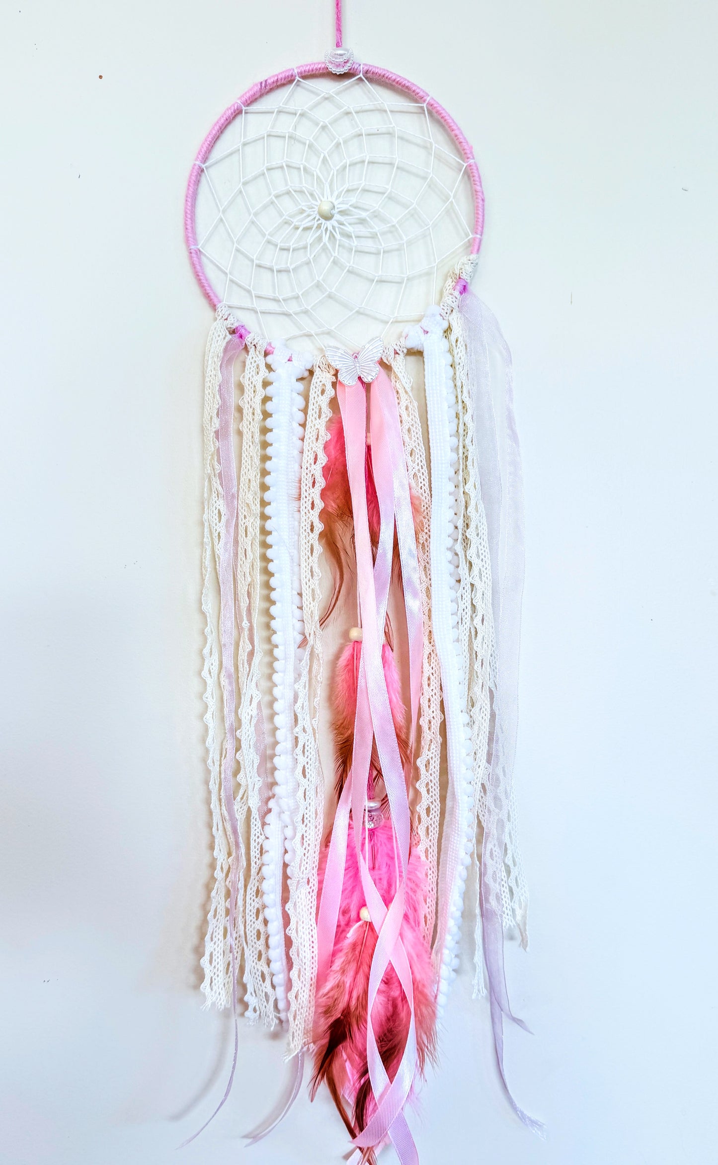 Pink dream catcher with a woven web, lace, and flowing ribbons in pastel tones, adding softness to any space.